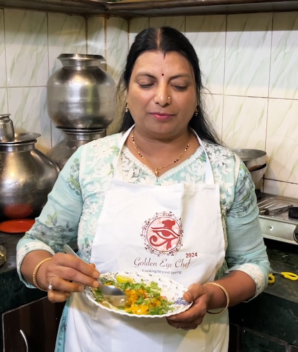 Pinky Mangal, a blind chef and participant of Golden Eye Chef, stands in a kitchen wearing a Golden Eye Chef 2024 apron, holding a plate of freshly prepared food and tasting it with a spoon.